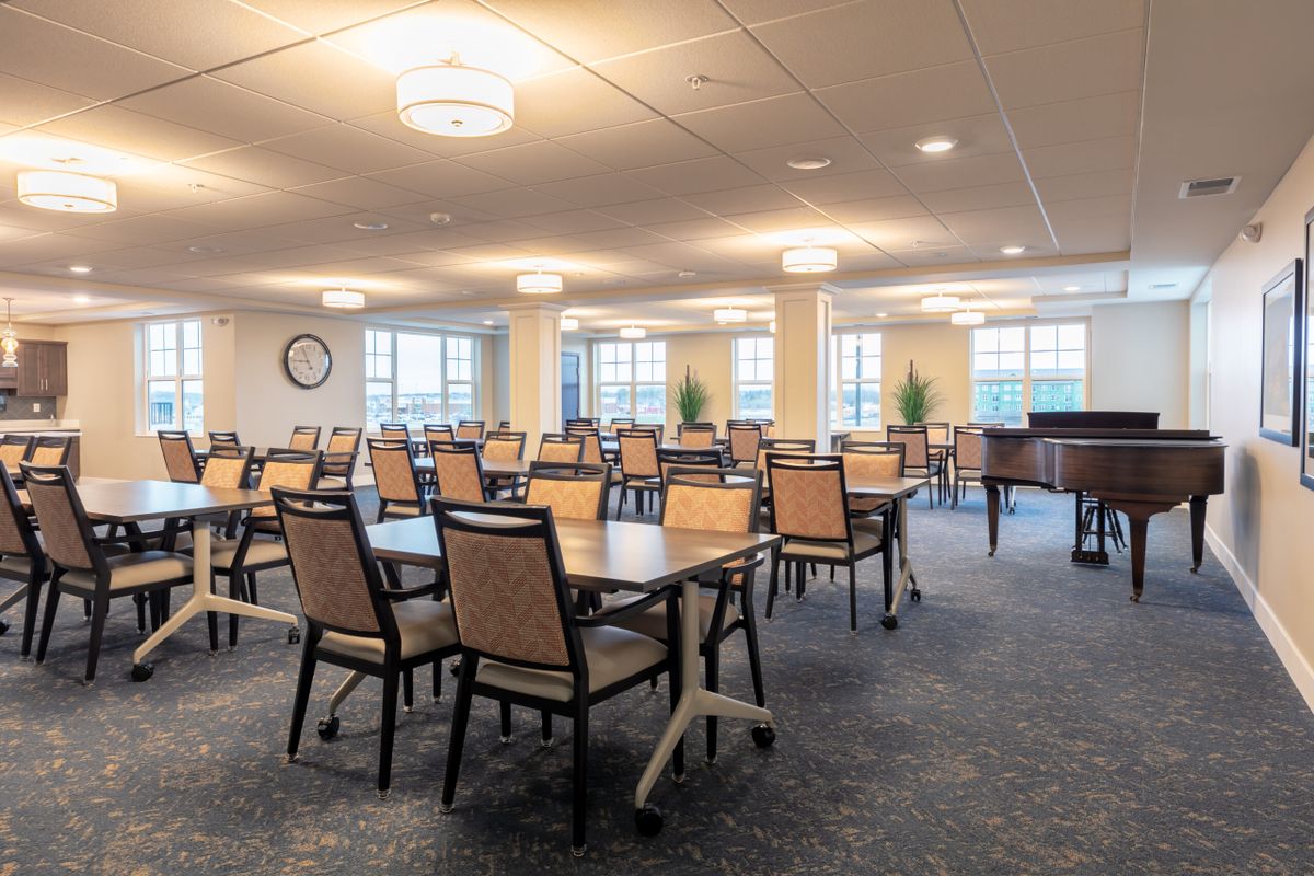A spacious dining room with multiple tables and chairs, well-lit by overhead lights and natural light from the windows.