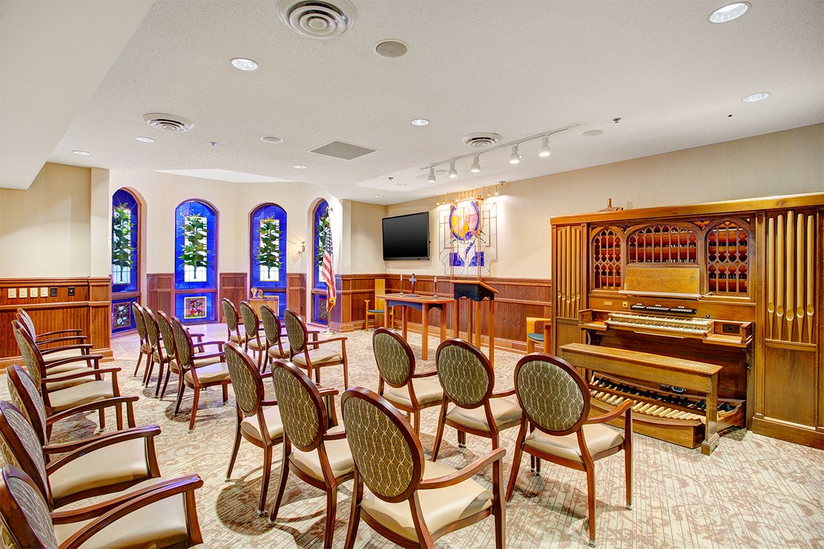 An interior view of a chapel with arranged chairs, stained glass windows, and an organ for worship.