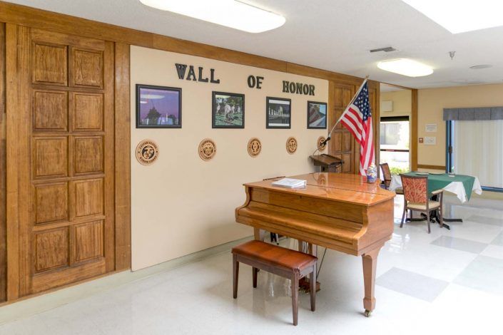 An interior room featuring a grand piano, an American flag, and a 'Wall of Honor' display with photos and decorations.