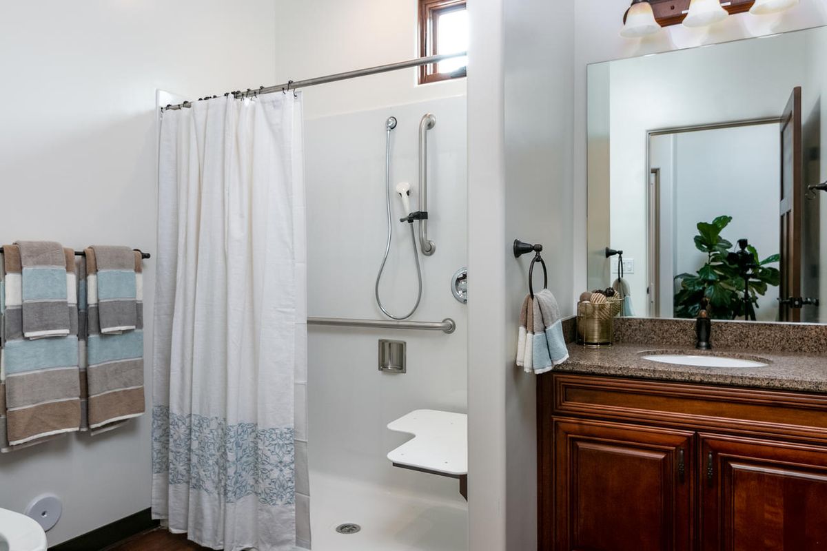 An accessible bathroom with a shower featuring grab bars and a foldable seat, alongside a vanity with a mirror.