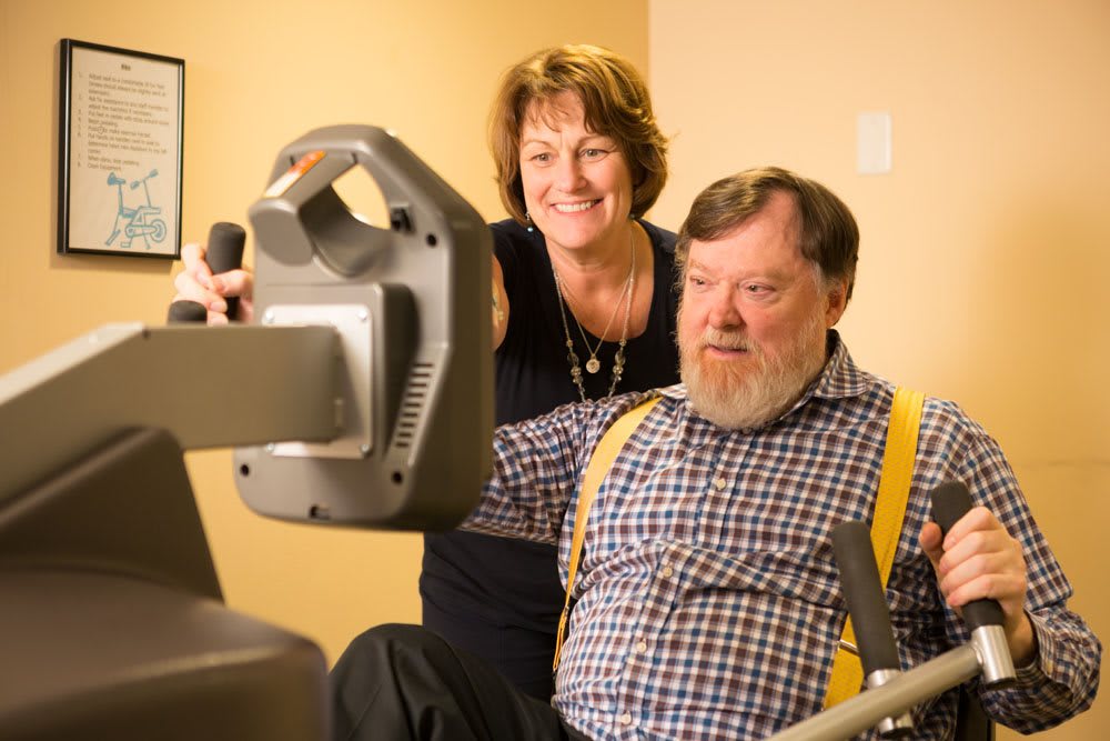 An elderly man receives assistance while using exercise equipment.