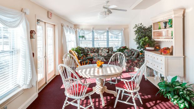 A cozy sunroom with a round table, chairs, and a floral-patterned couch surrounded by large windows letting in natural light.