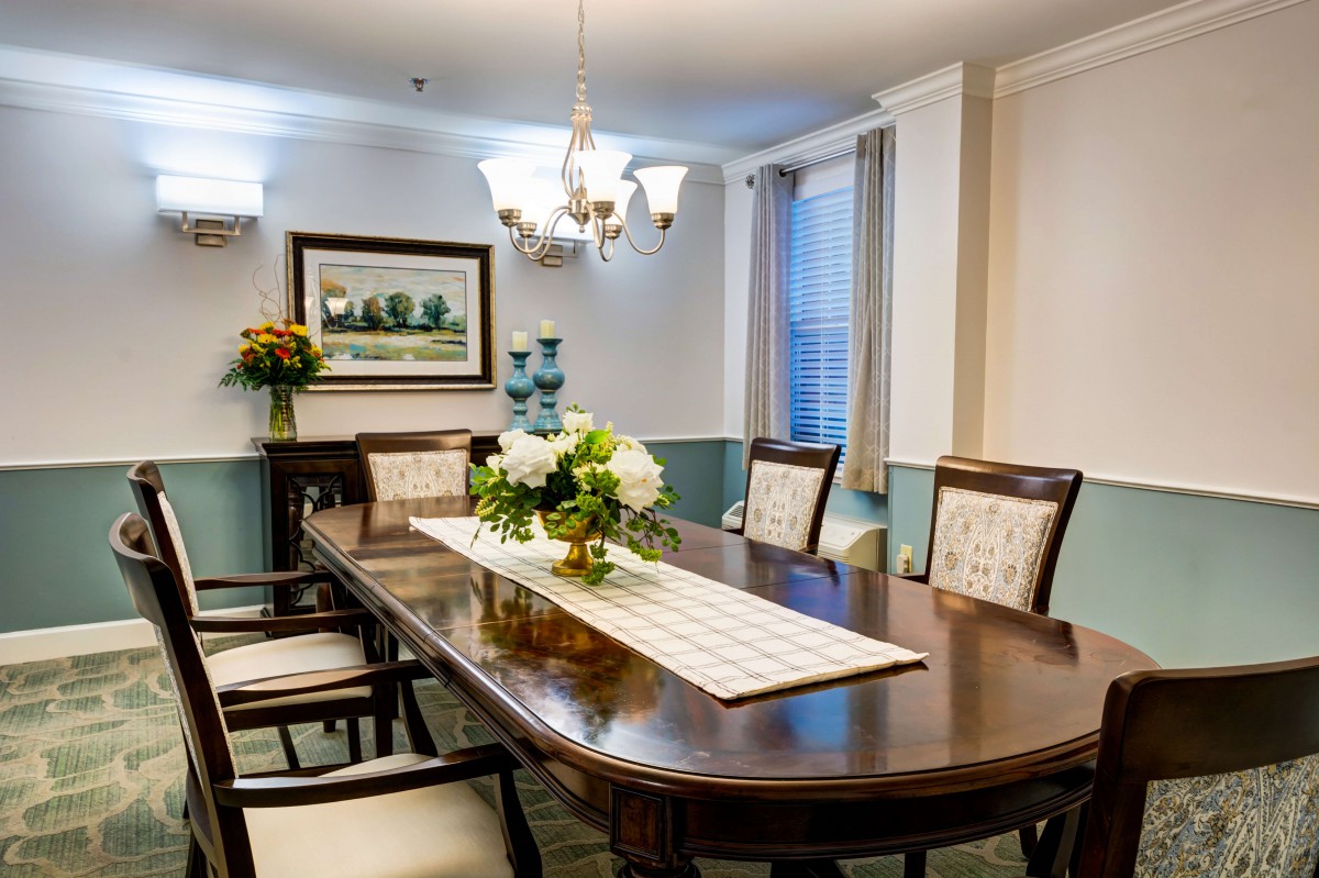 An elegantly decorated dining room with a central table, surrounded by chairs and adorned with a floral centerpiece under a chandelier.