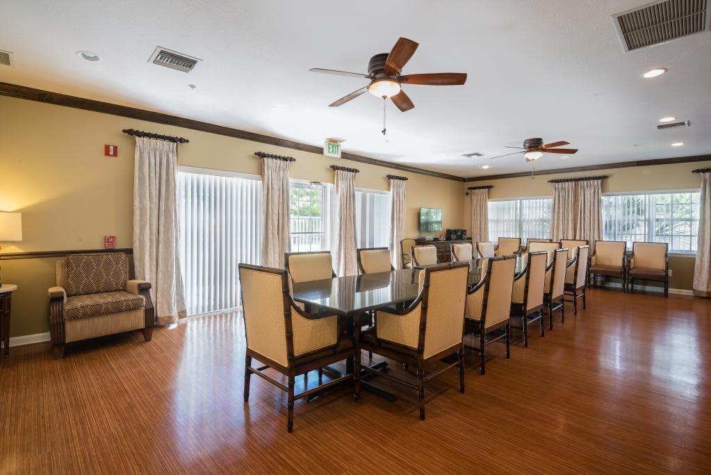 A formal dining room with a long table surrounded by chairs, featuring ceiling fans and natural light from large windows.