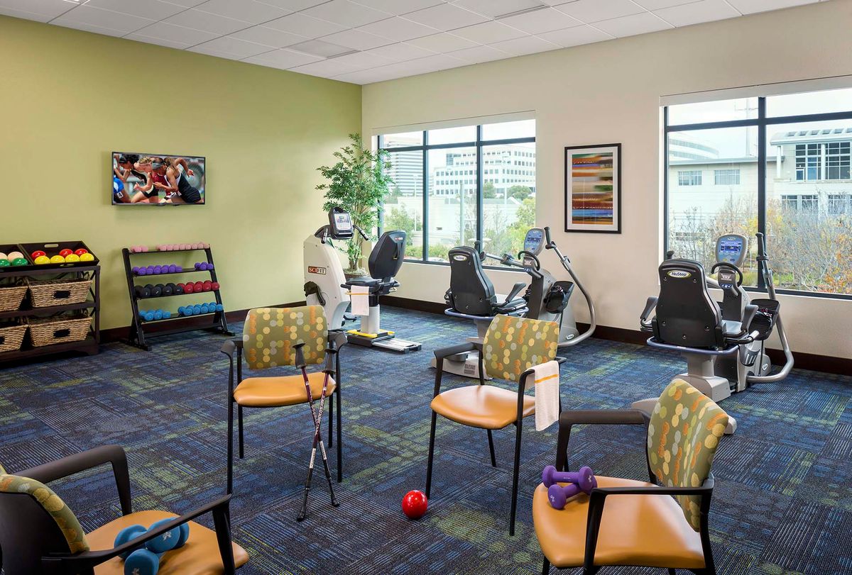 A well-lit gym room with exercise equipment and chairs arranged neatly.
