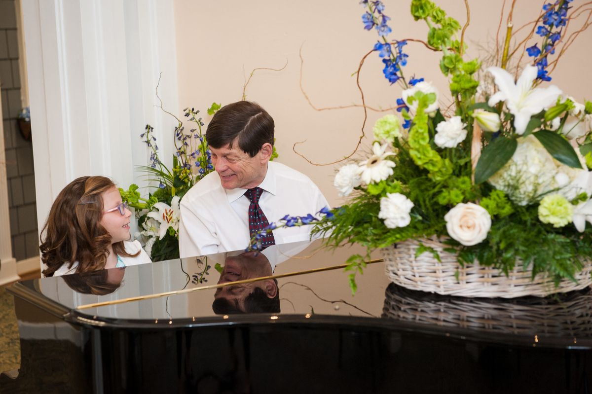 A man and a child smiling at each other while sitting at a piano, with a bouquet of flowers nearby.