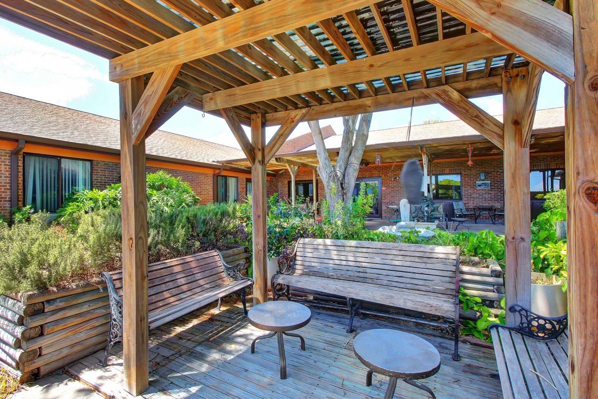 A charming outdoor space features a wooden pergola with benches and tables, surrounded by lush greenery.