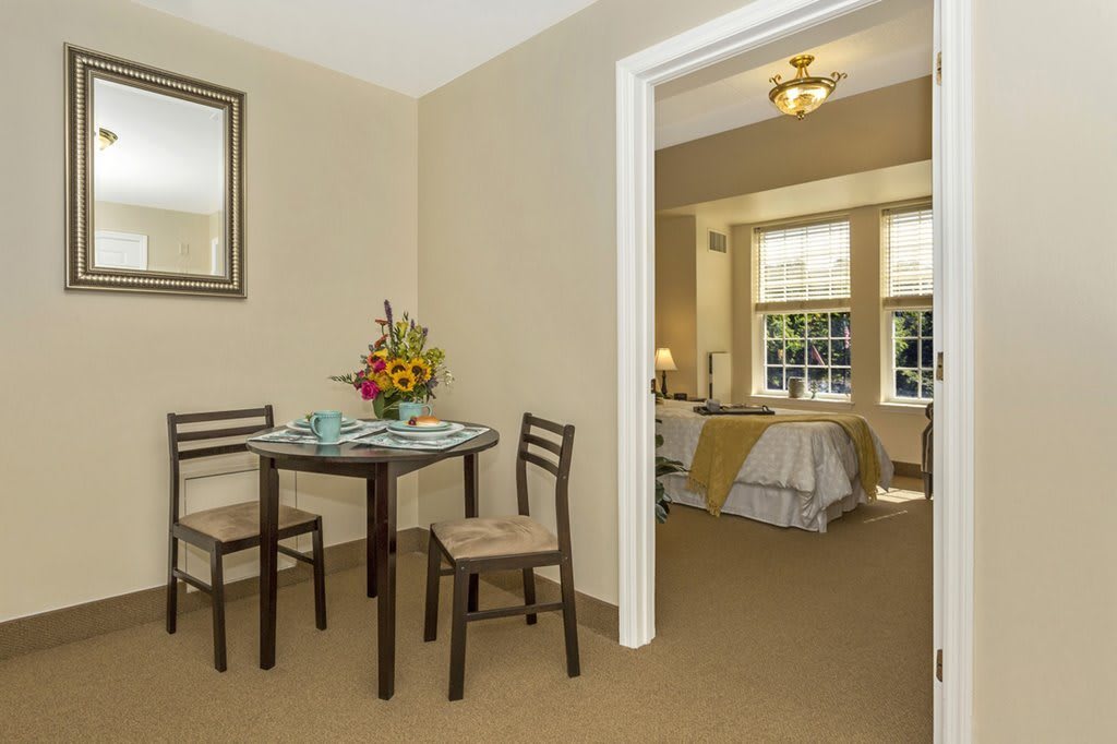 A cozy living space featuring a dining area with a table and chairs, leading to a bedroom with large windows.
