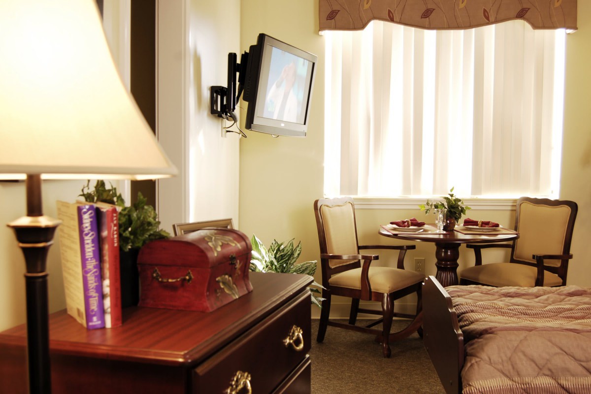 A cozy room featuring a television mounted on the wall, a wooden table with chairs, and a lamp on a dresser.