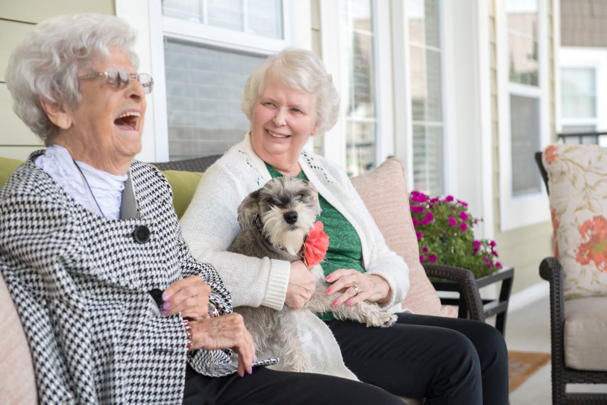 Two elderly women sitting outside, smiling and enjoying time with a small dog.