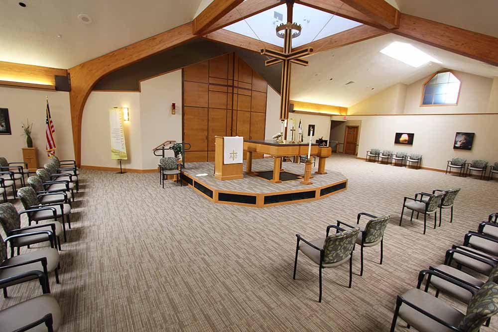 A peaceful chapel interior with seating arranged facing the altar and a prominent cross hanging from the ceiling.