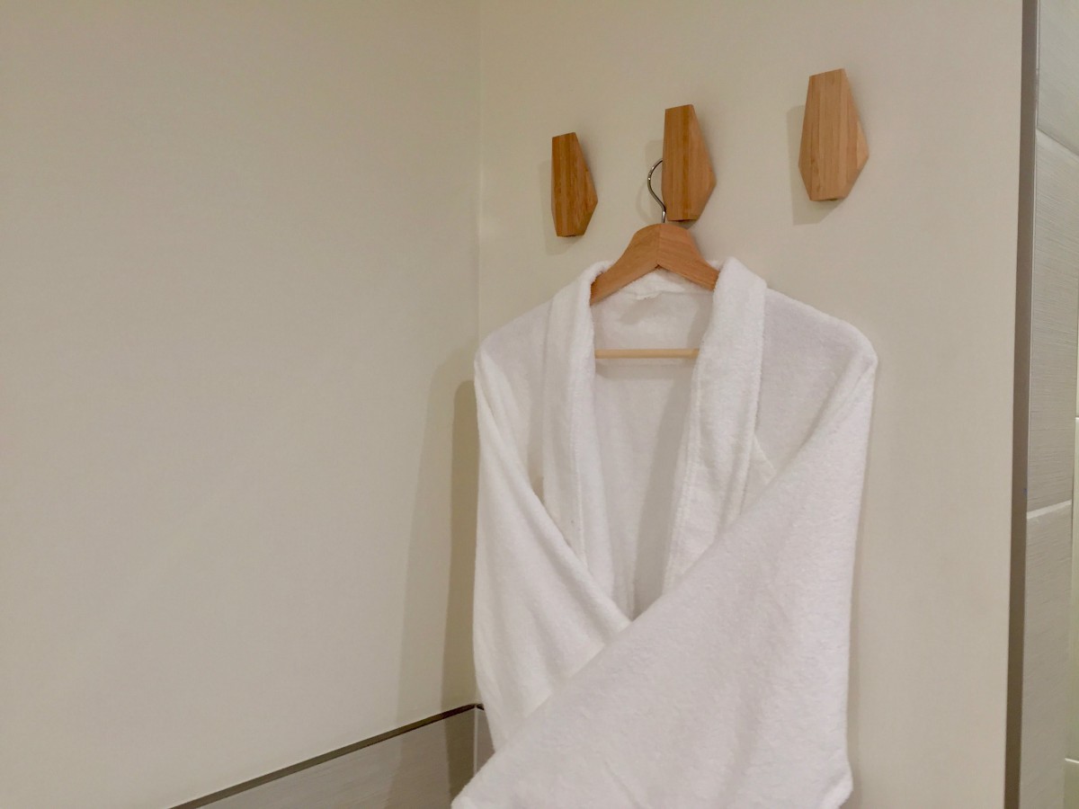 A white bathrobe hangs on a wooden hanger against a minimalist wall with wooden hooks.