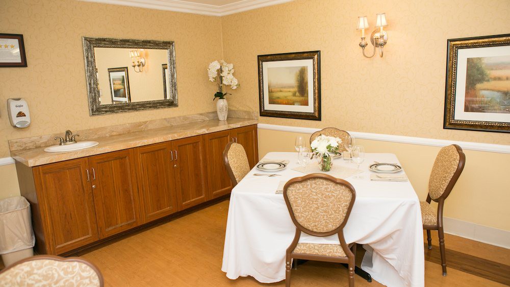 A warmly decorated dining room with a table set for two, featuring elegant chairs, a painting on the wall, and a large mirror above a sideboard.