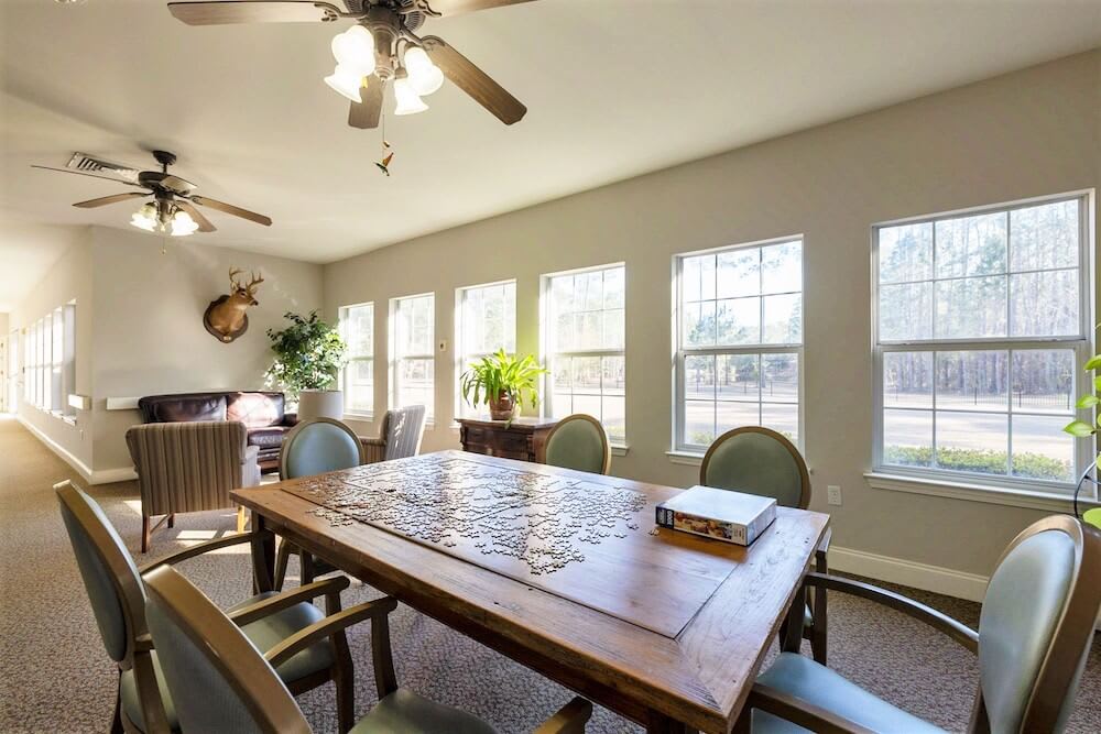 A well-lit dining room with a wooden table, surrounded by chairs, and illuminated by natural light through large windows.