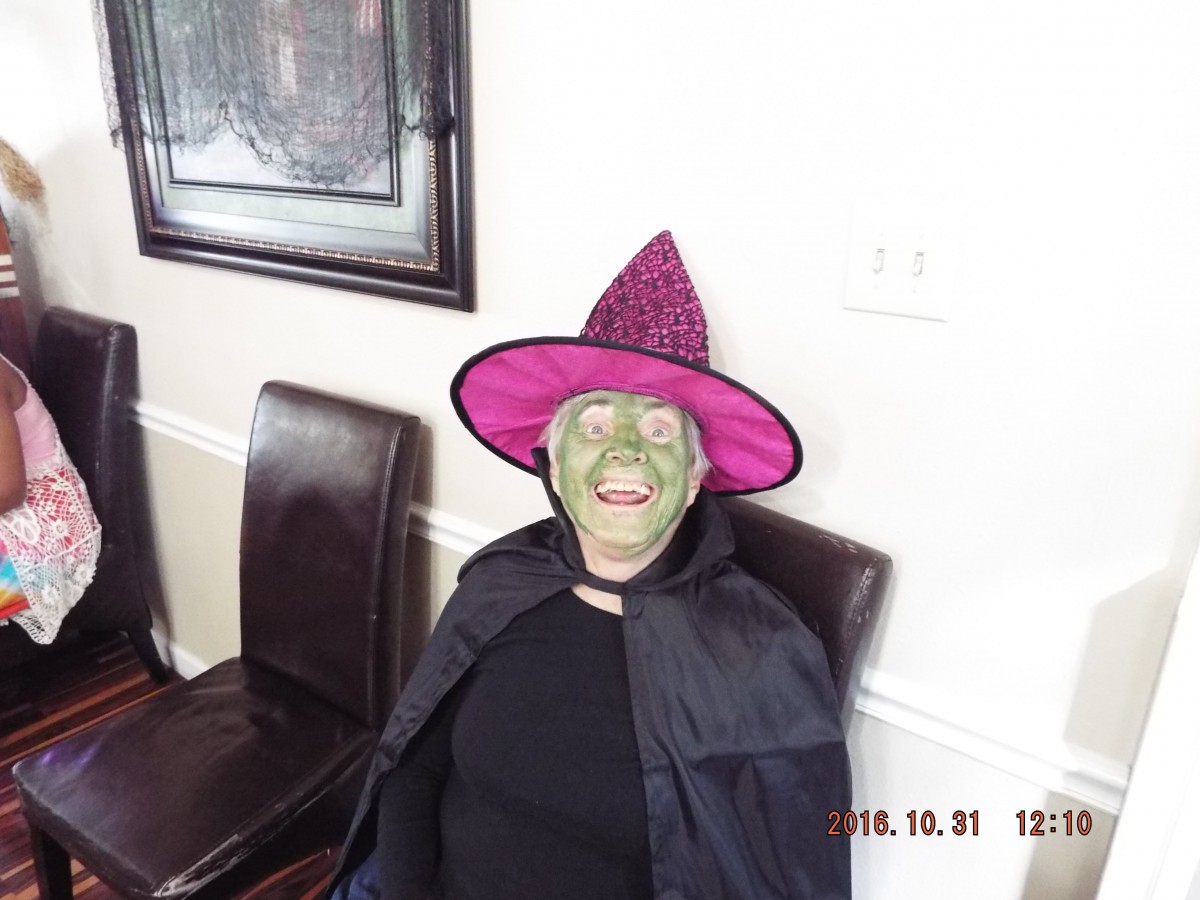 Person dressed as a witch with green face makeup and a pink hat, smiling indoors.