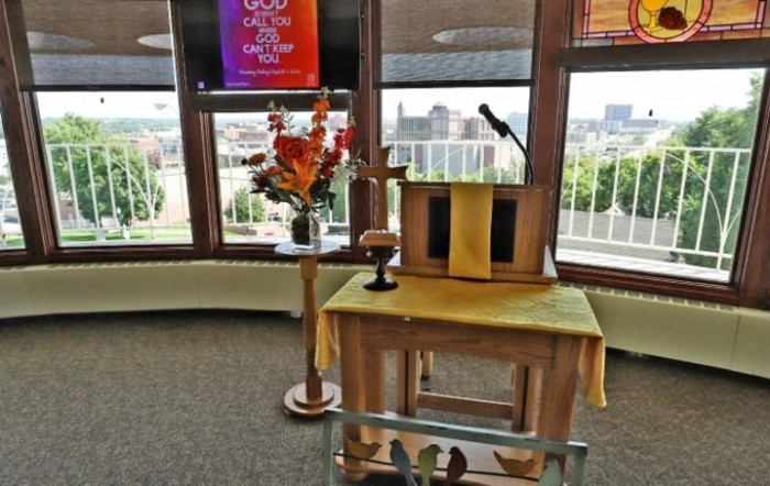 A church podium with a microphone and flower arrangement, set in front of large windows offering a scenic view.