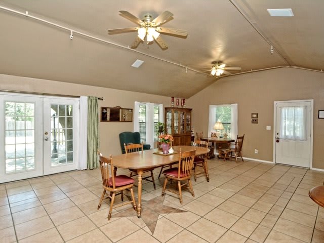 A spacious dining room with wooden furniture and a tiled floor, featuring ceiling fans and large windows.