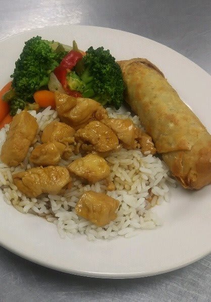 A plate featuring chicken over rice with a side of broccoli, red bell peppers, carrots, and a spring roll.