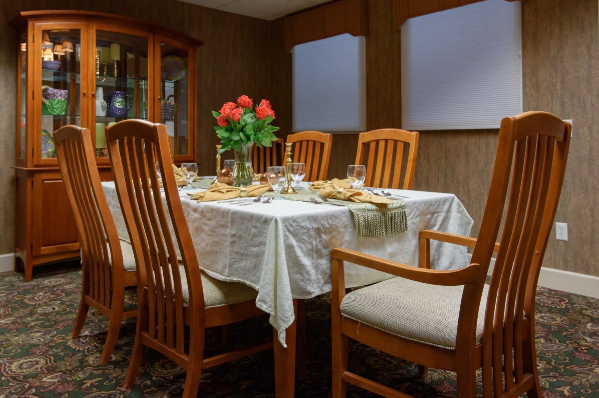 A dining room table set for a meal with wooden chairs and a vase of red roses as the centerpiece.