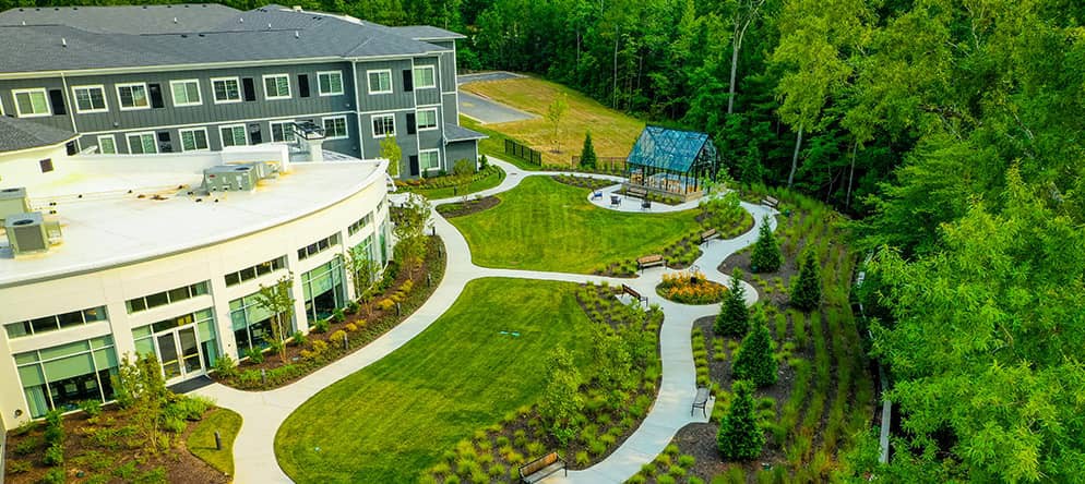 Aerial view of a landscaped garden with pathways, a building, and a small greenhouse surrounded by trees.