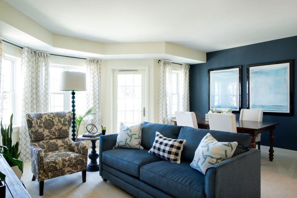 A cozy living room with a blue sofa, patterned armchair, and a bright dining area in the background.