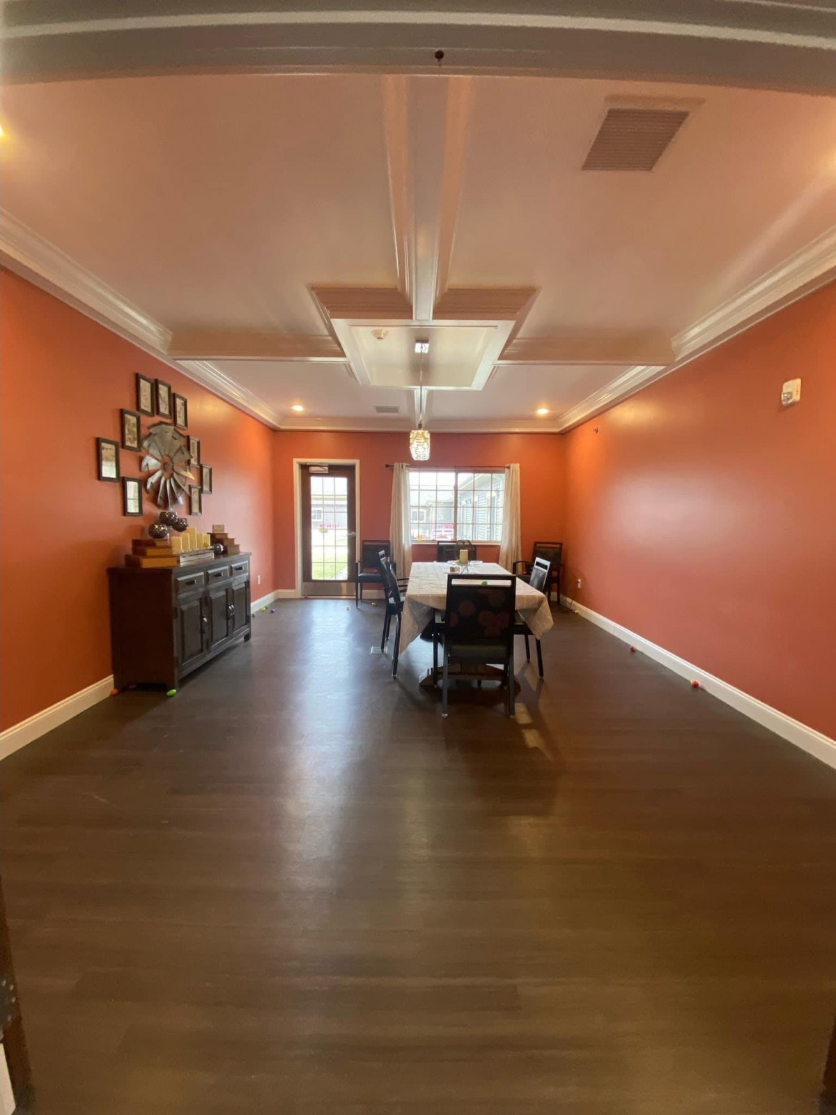 A spacious dining room with vibrant orange walls, featuring a central table and chairs, wood flooring, and decorative items on a sideboard.