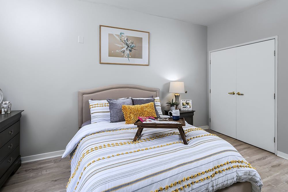 A cozy bedroom with a neatly made bed, decorative pillows, and a tray on the duvet. The room features a nightstand with a lamp and wall art above the headboard.