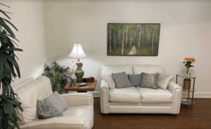 A cozy living room featuring white sofas, a table lamp, and a nature-themed painting.