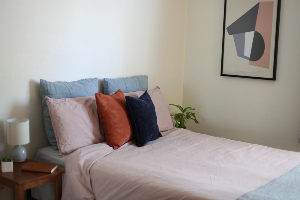 A neatly arranged bedroom with a bed adorned with colorful pillows, accompanied by artwork and a plant.