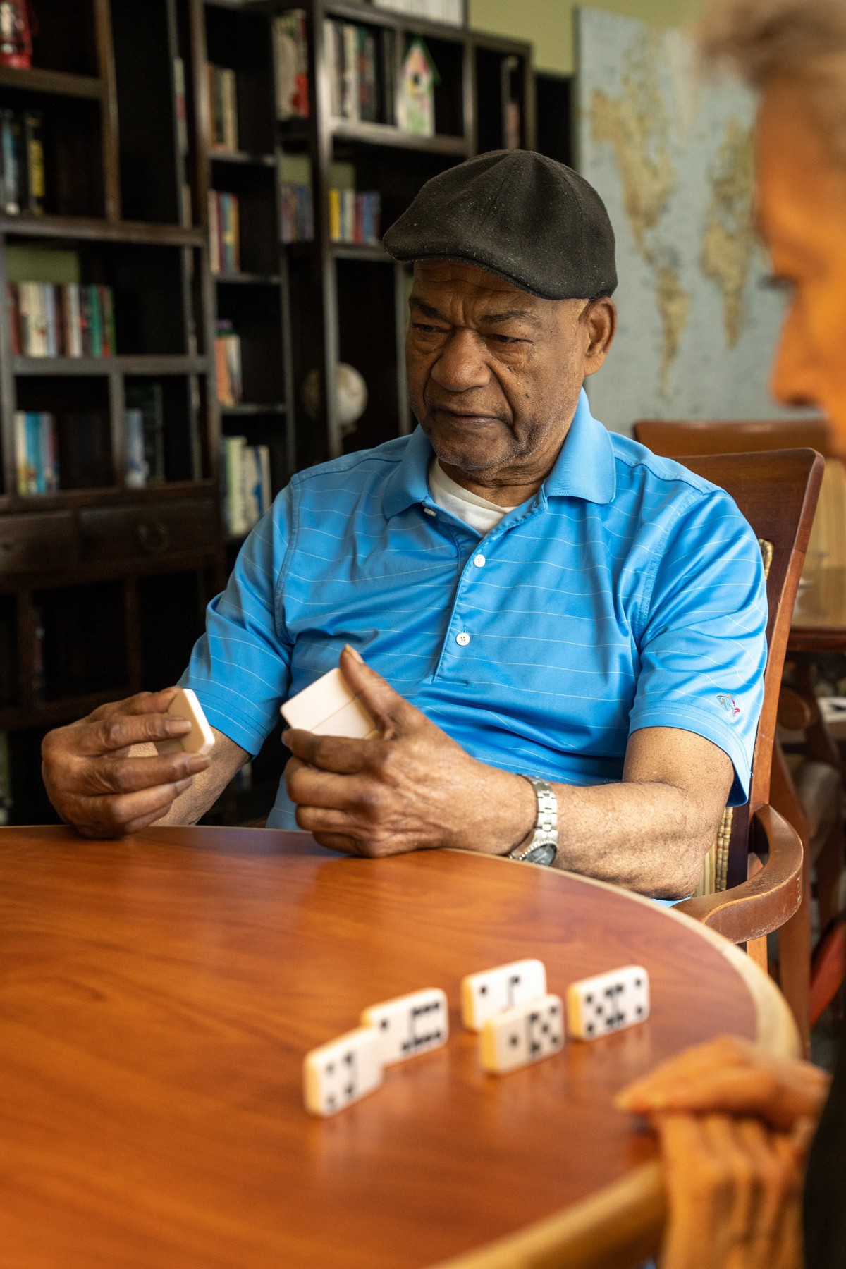 An elderly man in a blue shirt and cap playing cards at a table with dominoes.