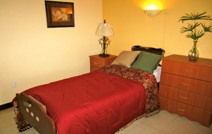 A cozy bedroom with a single bed, red bedding, and a lamp on the nightstand.