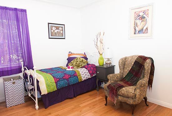 A cozy bedroom with colorful bedding, an armchair draped with a blanket, and decorative wall art.