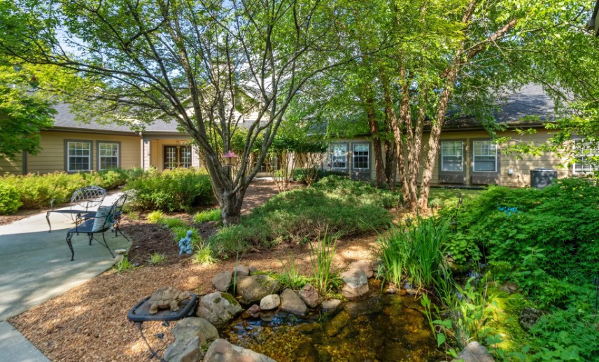 A serene garden with lush greenery and a small pond, surrounded by buildings.