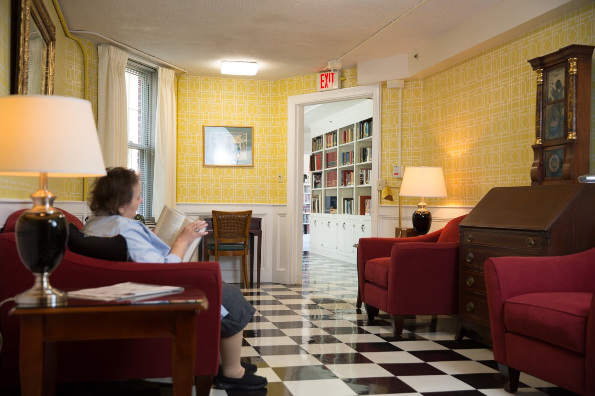 A cozy living room with yellow walls, red chairs, and a checkerboard floor. A person is seated, reading a book.