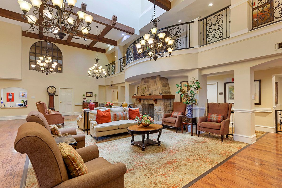 A spacious and luxurious living room with elegant chandeliers, wooden ceiling beams, a grand fireplace, and comfortable seating arranged around a large carpet.