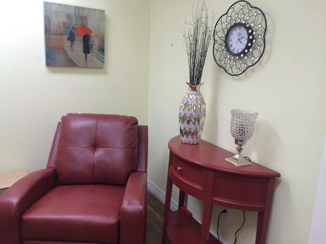 A cozy corner featuring a red armchair and matching side table, accented with a vase, wall art, and a decorative clock.