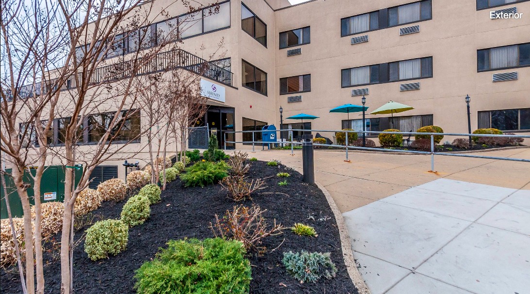 Exterior view of a building entrance with landscaping and a seating area with umbrellas.