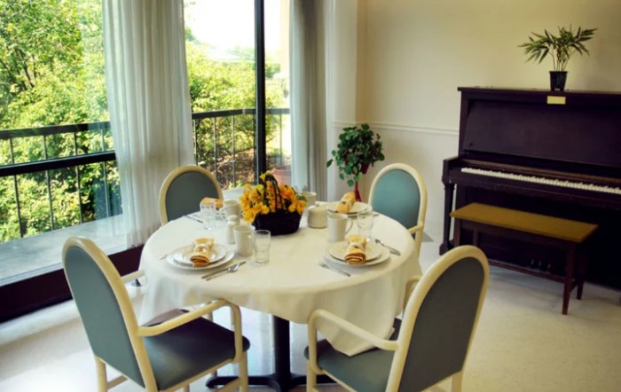 A bright and inviting dining area with a round table set for four, featuring a centerpiece of sunflowers and a piano in the background.
