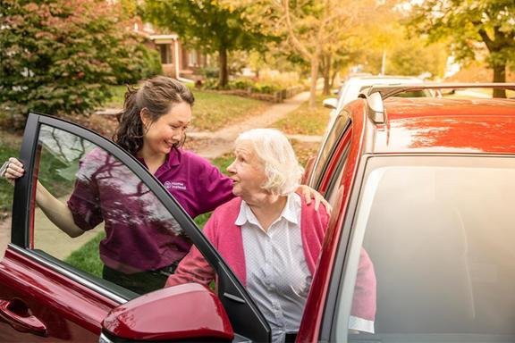 A young woman assists an elderly woman getting out of a car in an outdoor setting.