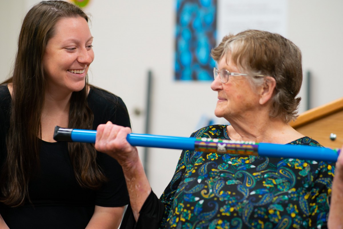 An elderly woman lifting a blue bar while being encouraged by a younger woman.