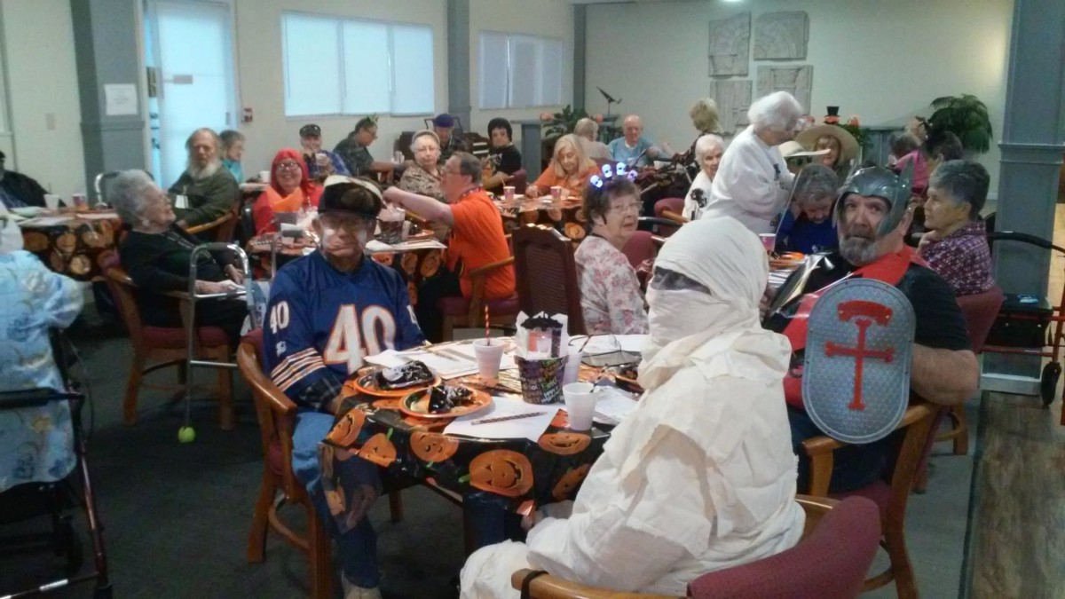 A lively Halloween celebration with attendees in various costumes, including a mummy and a person in a sports jersey, seated at decorated tables.