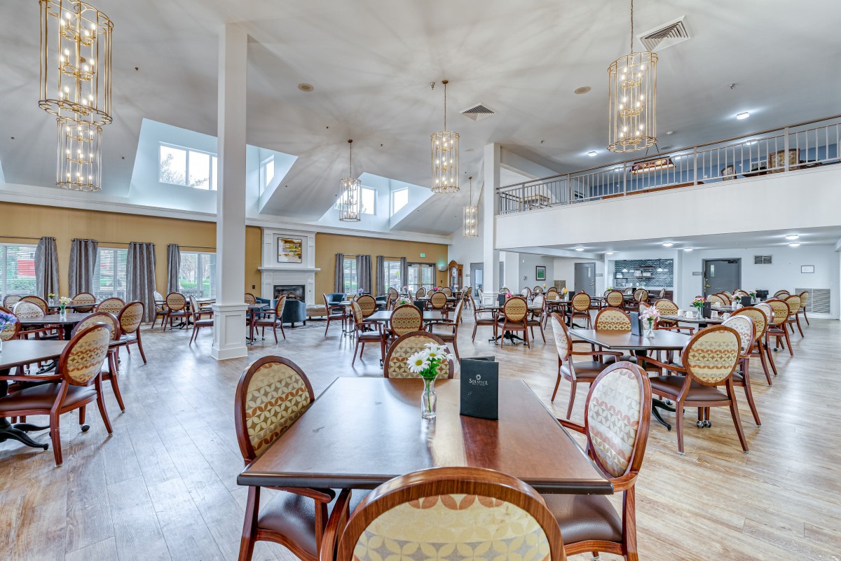 A spacious and elegantly designed restaurant interior with multiple dining tables and chairs under a high ceiling adorned with chandeliers.