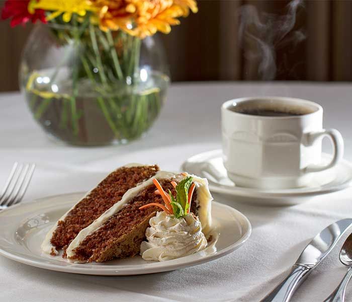 A slice of carrot cake with cream cheese frosting and a cup of coffee on a table decorated with flowers.