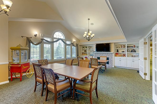 A spacious dining area with a large table, surrounded by chairs, featuring a chandelier and large windows, with shelves and a television in the background.