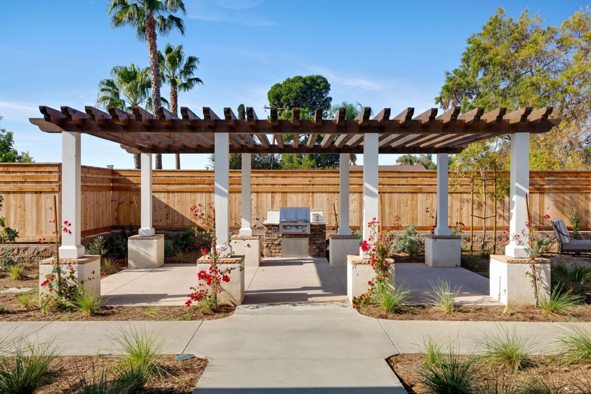 A sunny backyard featuring a pergola with outdoor seating surrounded by lush landscaping and a wooden fence.