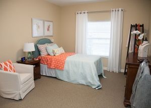 A cozy bedroom with a neatly made bed, adorned with pastel bedding and decorative pillows, alongside two framed artworks above the headboard.
