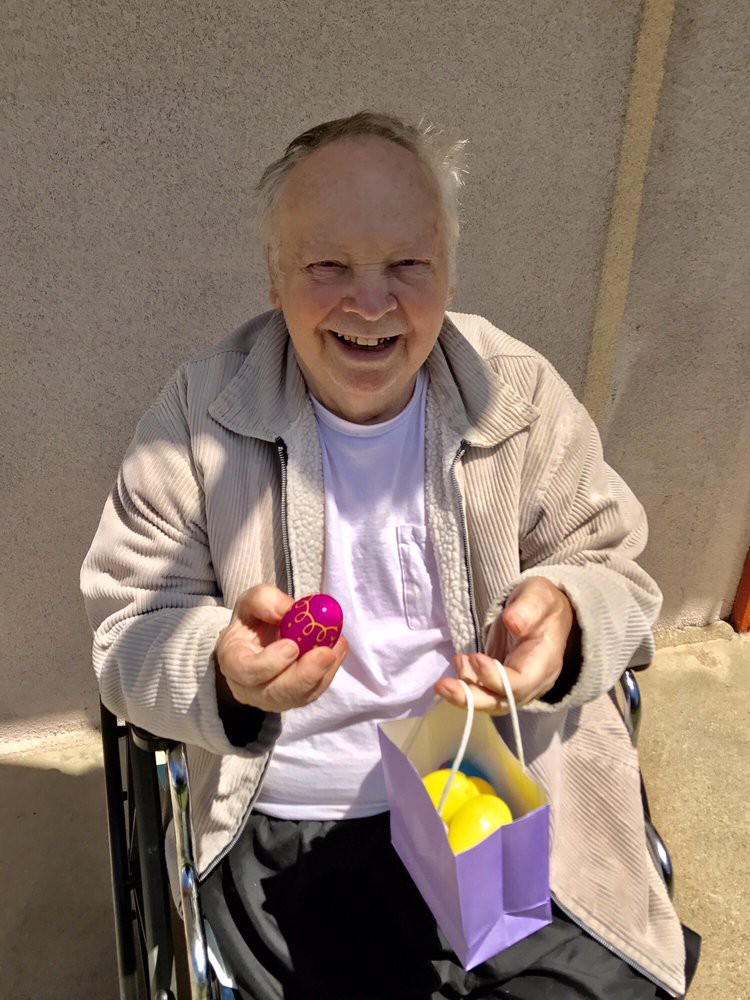 An elderly man smiling while holding a colorful egg and a gift bag outdoors.