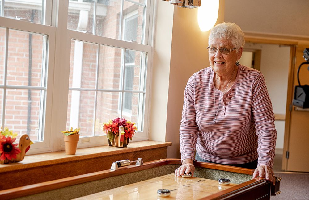 An elderly woman enjoys a game of shuffleboard indoors by the window, with flowers in the background.