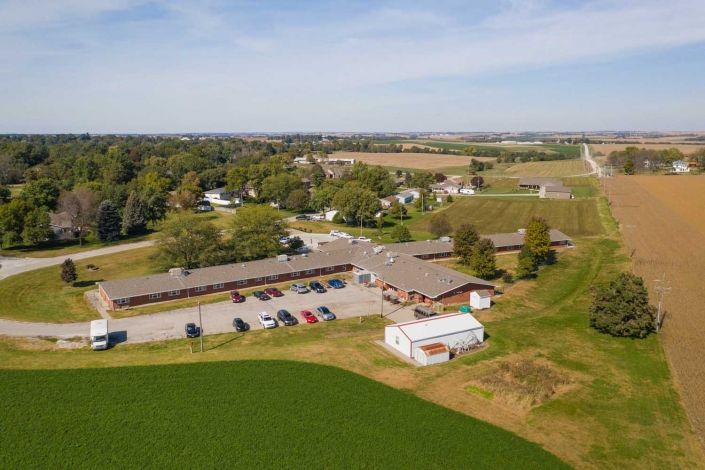 An aerial view of a rural landscape featuring farmland, a parking lot, and several buildings surrounded by greenery.