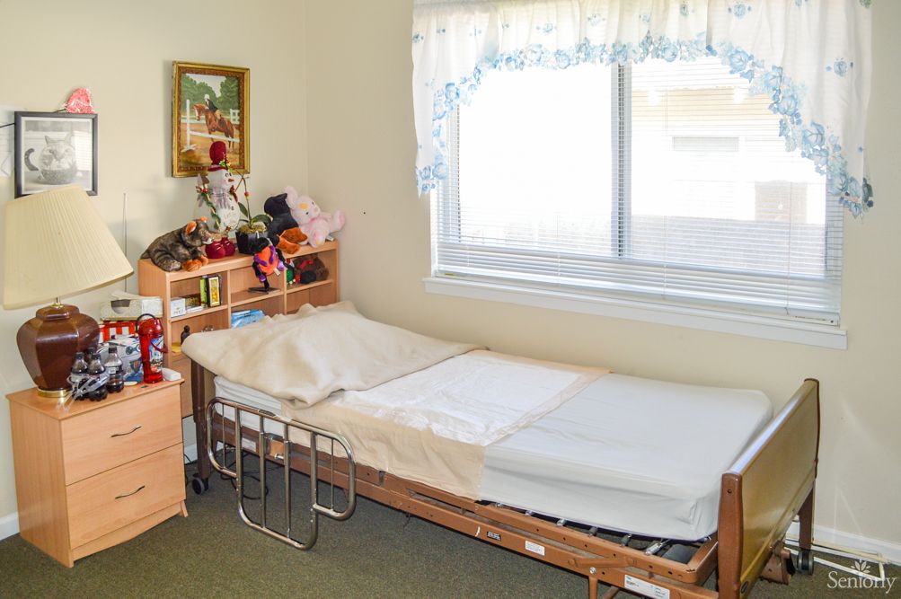 A cozy bedroom featuring a hospital bed, a window with lace curtains, and a neatly organized bookshelf filled with books and decorations.